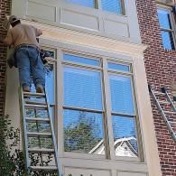 A person on a ladder repairs the trim around large windows on a multi-story brick house.