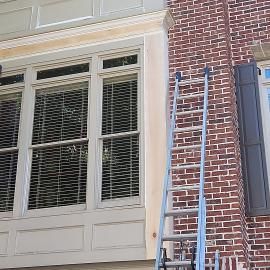 An extension ladder leans against a brick wall next to a large window with wooden trim being prepped for painting.