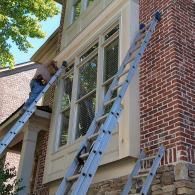 A person climbs a ladder leaned against the exterior of a brick house to reach a window.