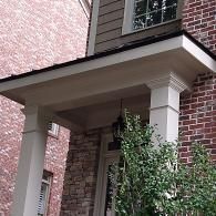 A close-up of a house entrance featuring white porch columns, a dark roof overhang, stone siding, and a hanging lantern.