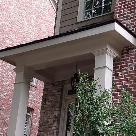 A white wooden entryway overhang supported by square columns against a brick and stone house exterior.