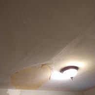 A damaged ceiling with a large patch of peeling, discolored paint next to an illuminated ceiling light fixture.