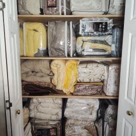 A closet with white shelving filled with neatly stacked, folded bedding and blankets stored in clear plastic bags.