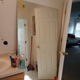 A bathroom vanity with toiletries reflected in a full-length mirror, showing an open bedroom door and a red broom.