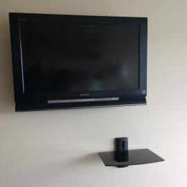 A flat-screen television mounted on a plain wall above a small, black, glass shelf.