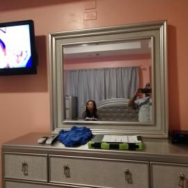 A bedroom dresser with a large silver-framed mirror reflects a person taking a photo, with a TV on the pink wall nearby.