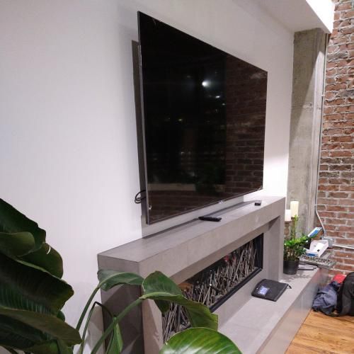 A large flat-screen TV mounted on a white wall above a long, concrete fireplace mantel next to a brick wall.