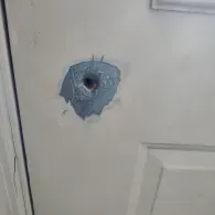 A white door with a circular hole, surrounded by splintered material and chipped paint, suggesting a bullet impact.