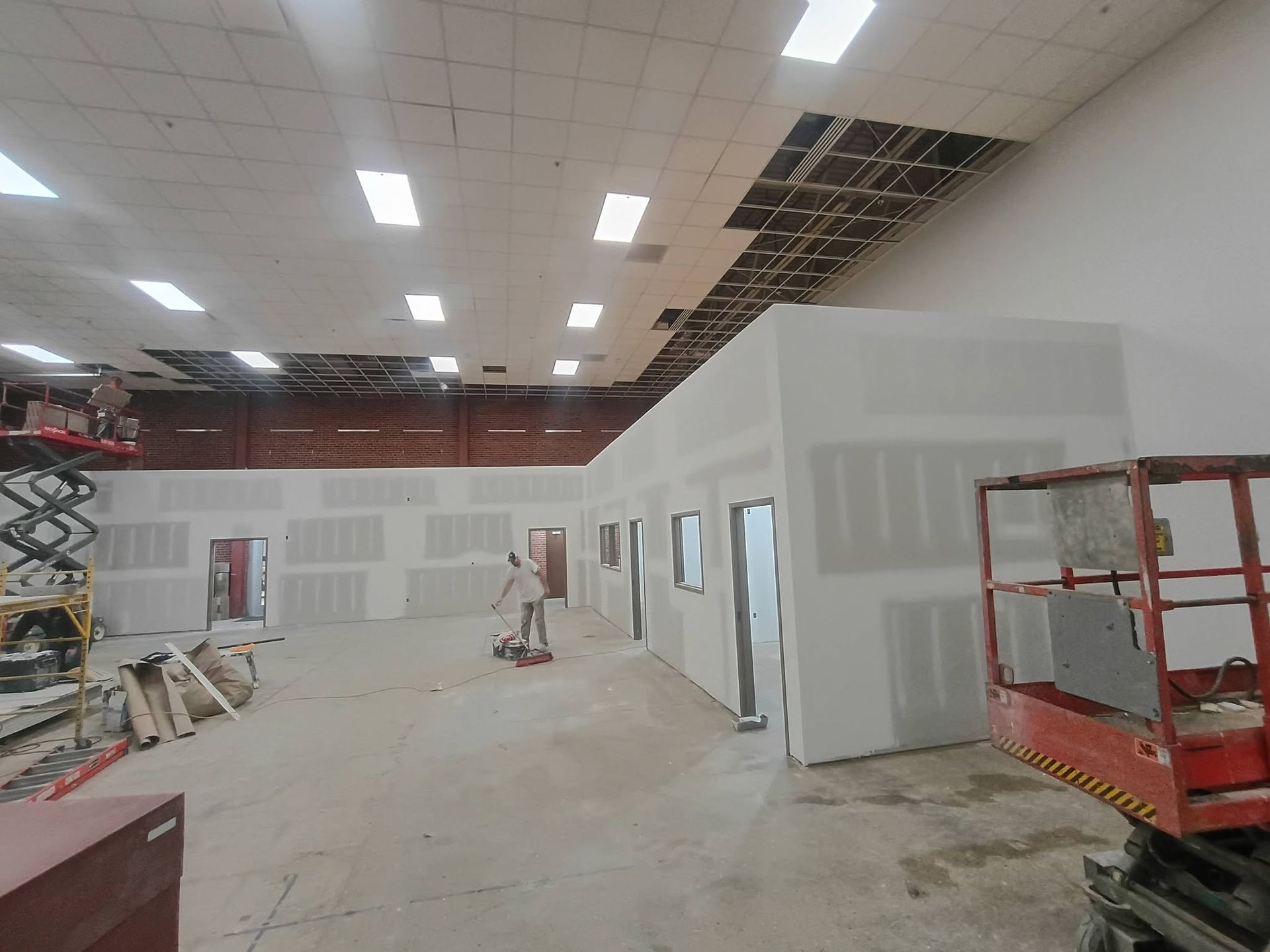 Construction site interior with drywall, open ceiling tiles, and red scaffolding under bright lights