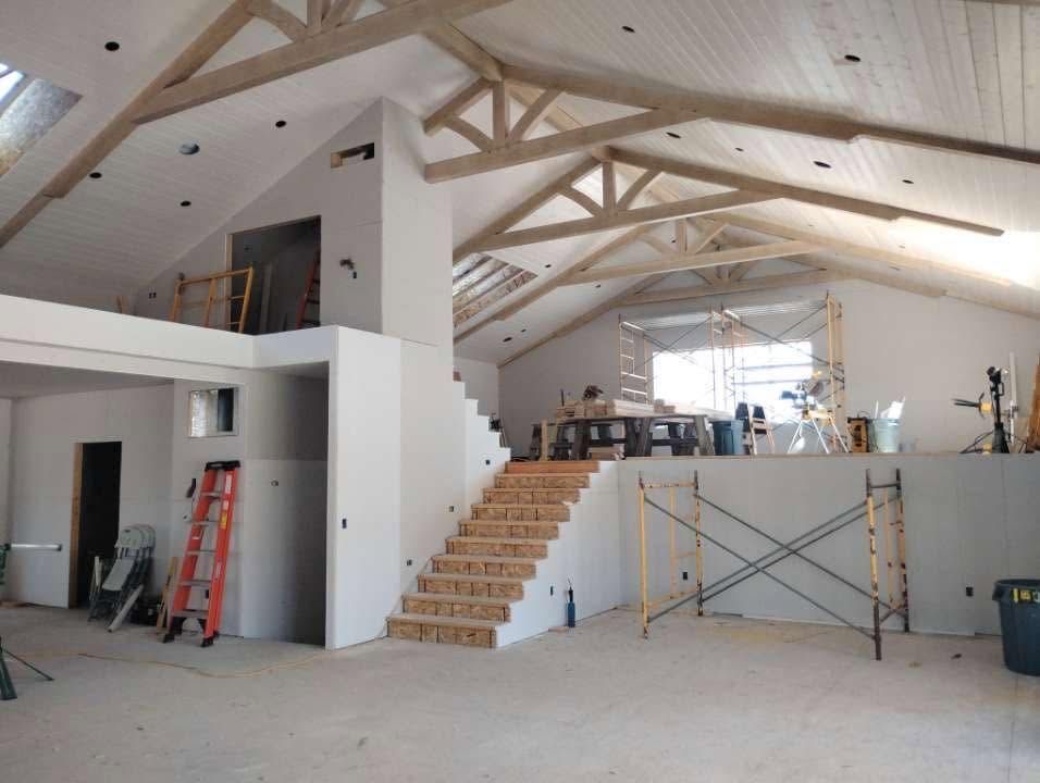 Unfinished room with vaulted ceiling, exposed beams, stairs, and construction equipment inside.