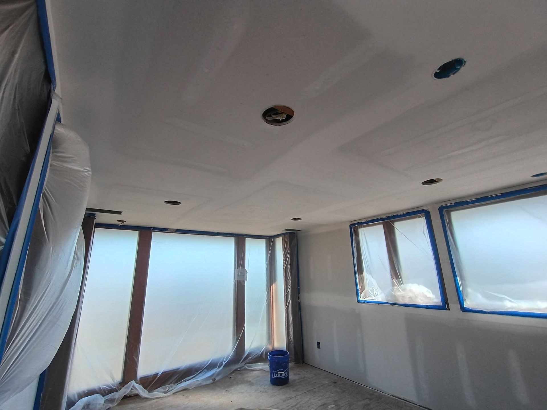 Empty room under renovation with white walls, large windows, plastic-covered surfaces, and a blue bucket on the floor