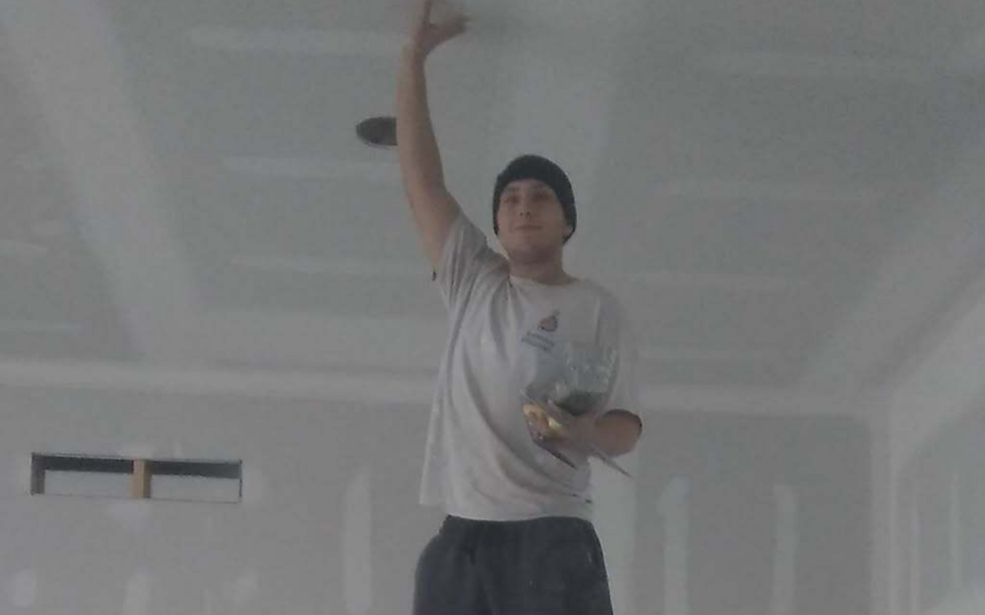 Person in a white shirt and black beanie stands in an unfinished room, reaching up with one arm.