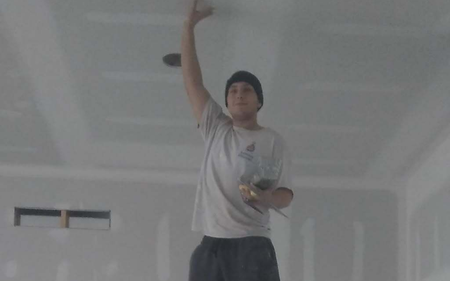 Person in a white shirt and black beanie stands in an unfinished room, reaching up with one arm.