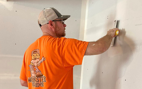 Worker in orange shirt smoothing drywall with a trowel in a partially finished room