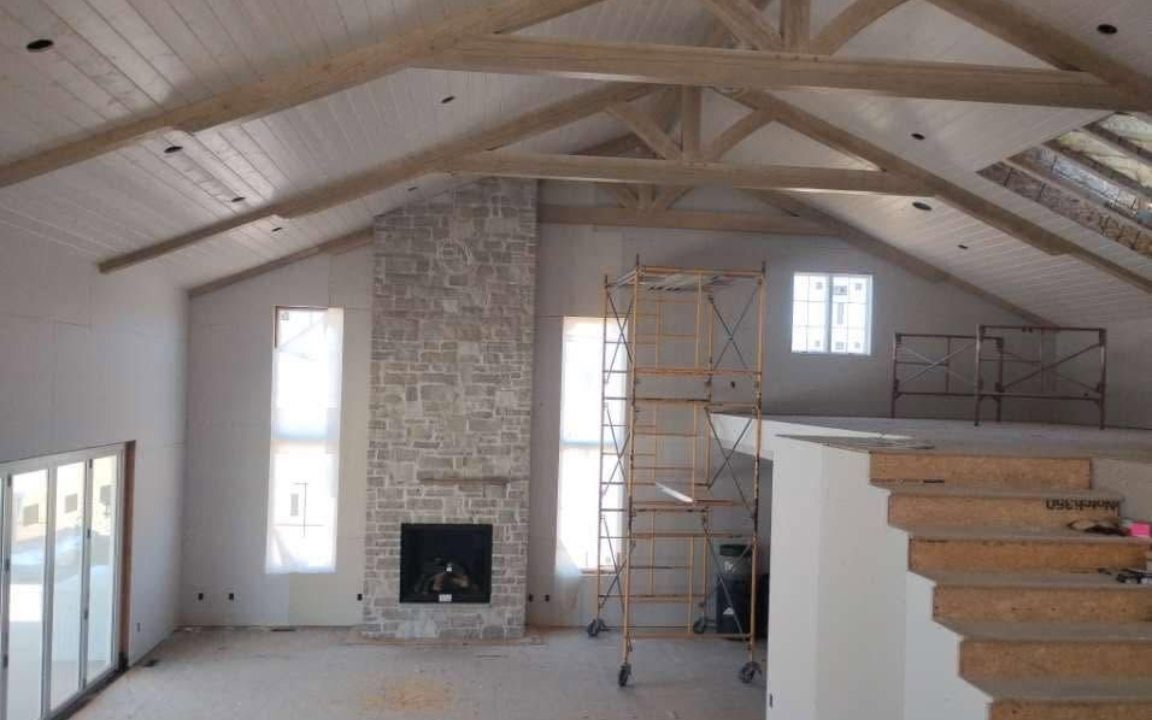 Bright unfinished living room with vaulted wood beams, stone fireplace, and staircase under construction