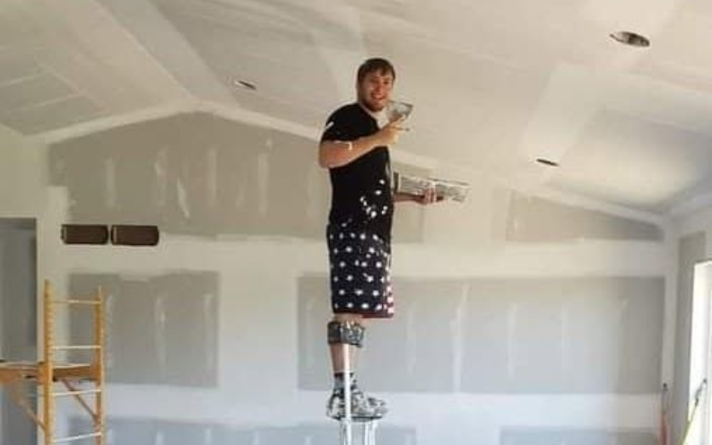 Person standing on stilts, applying drywall mud to a wall in a room under renovation.