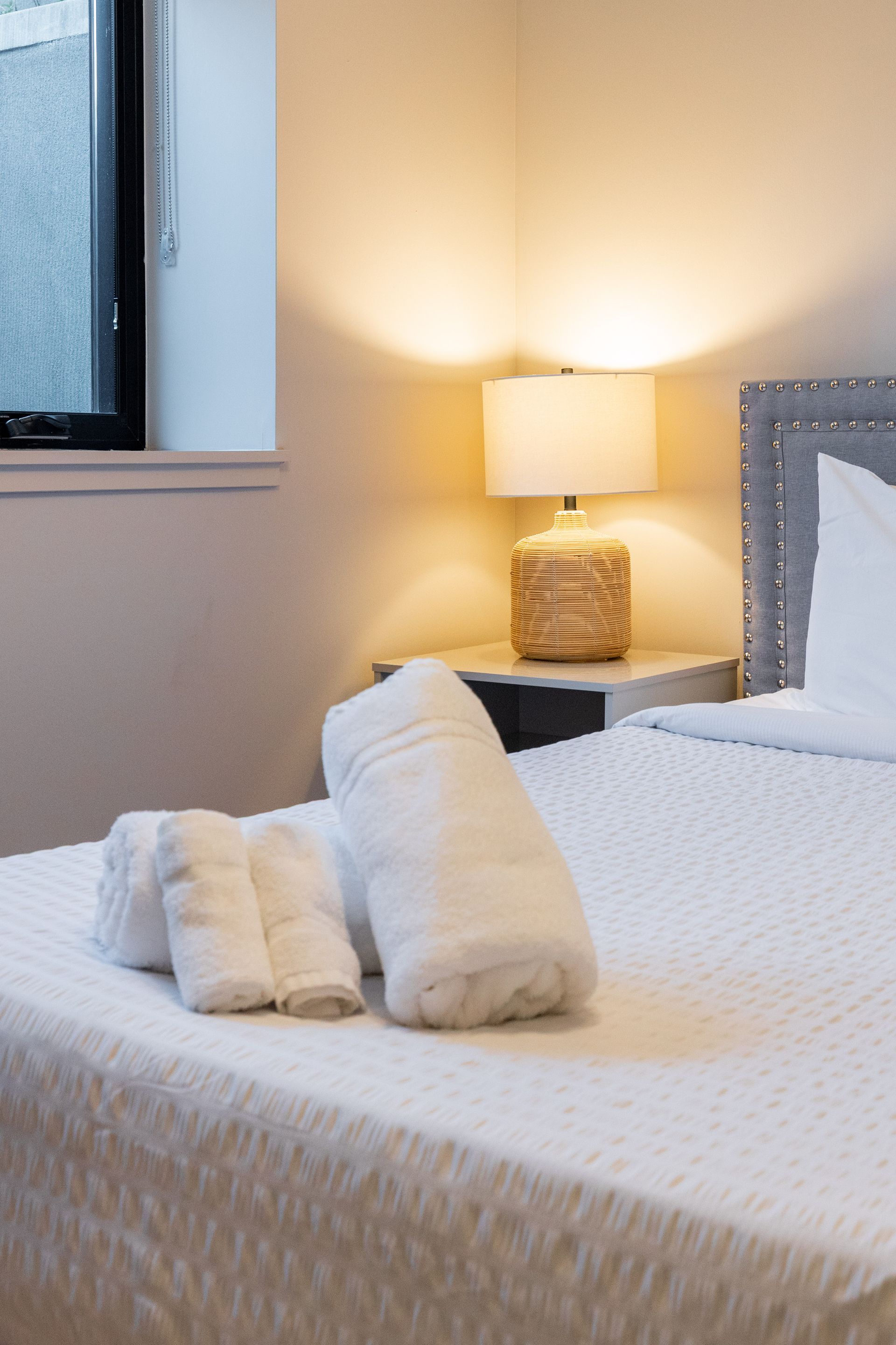 Neatly made bed with white bedding and towels beside a lit bedside lamp in a cozy bedroom.