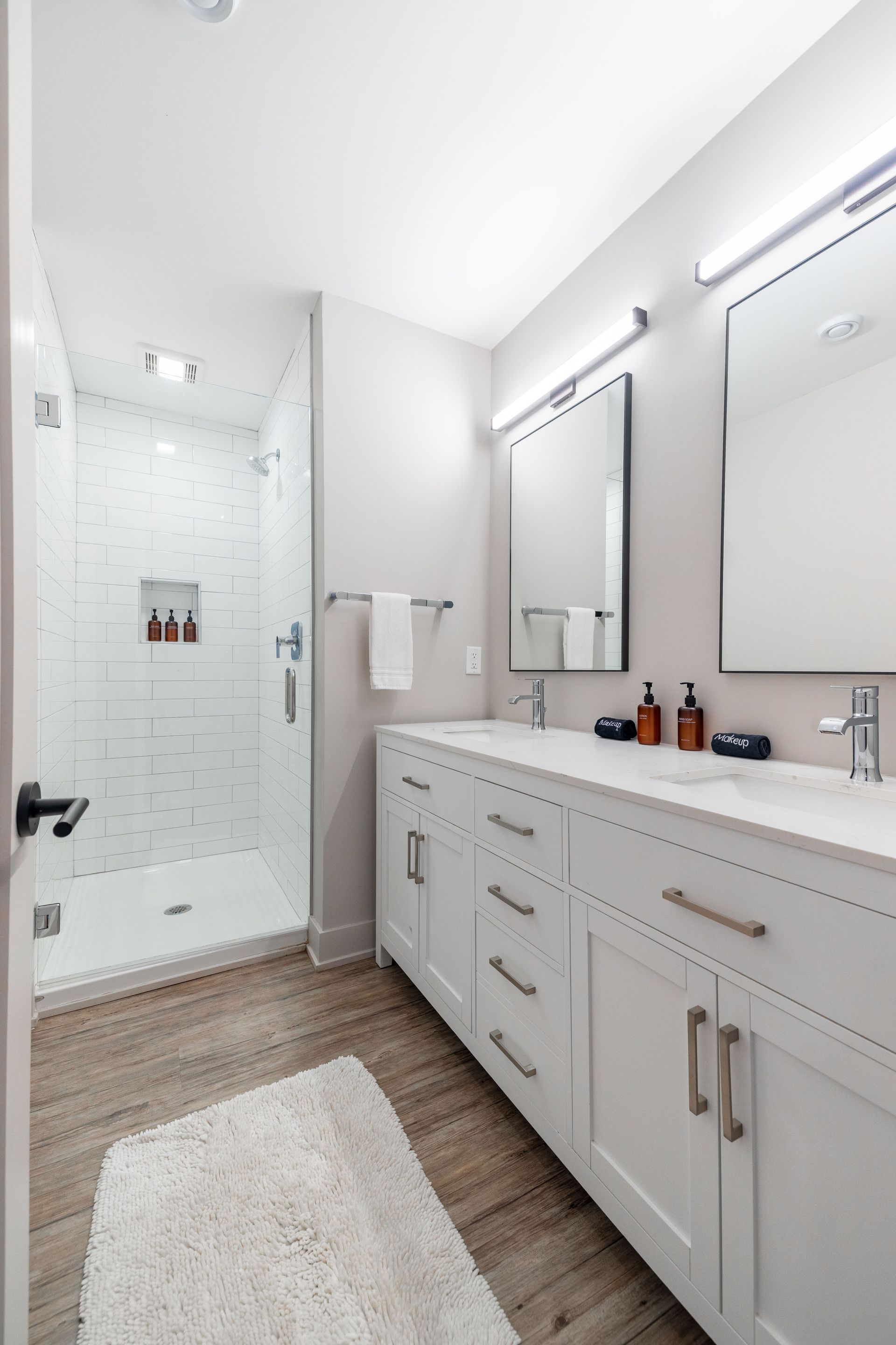 Bright white bathroom with double vanity, mirrors, glass shower, and beige rug on wood-look floor