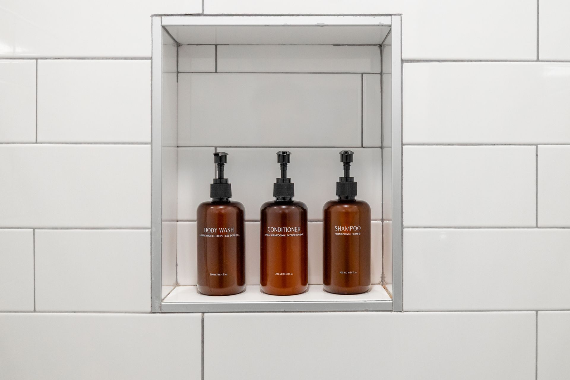 Three amber pump bottles on a white tiled recessed shelf