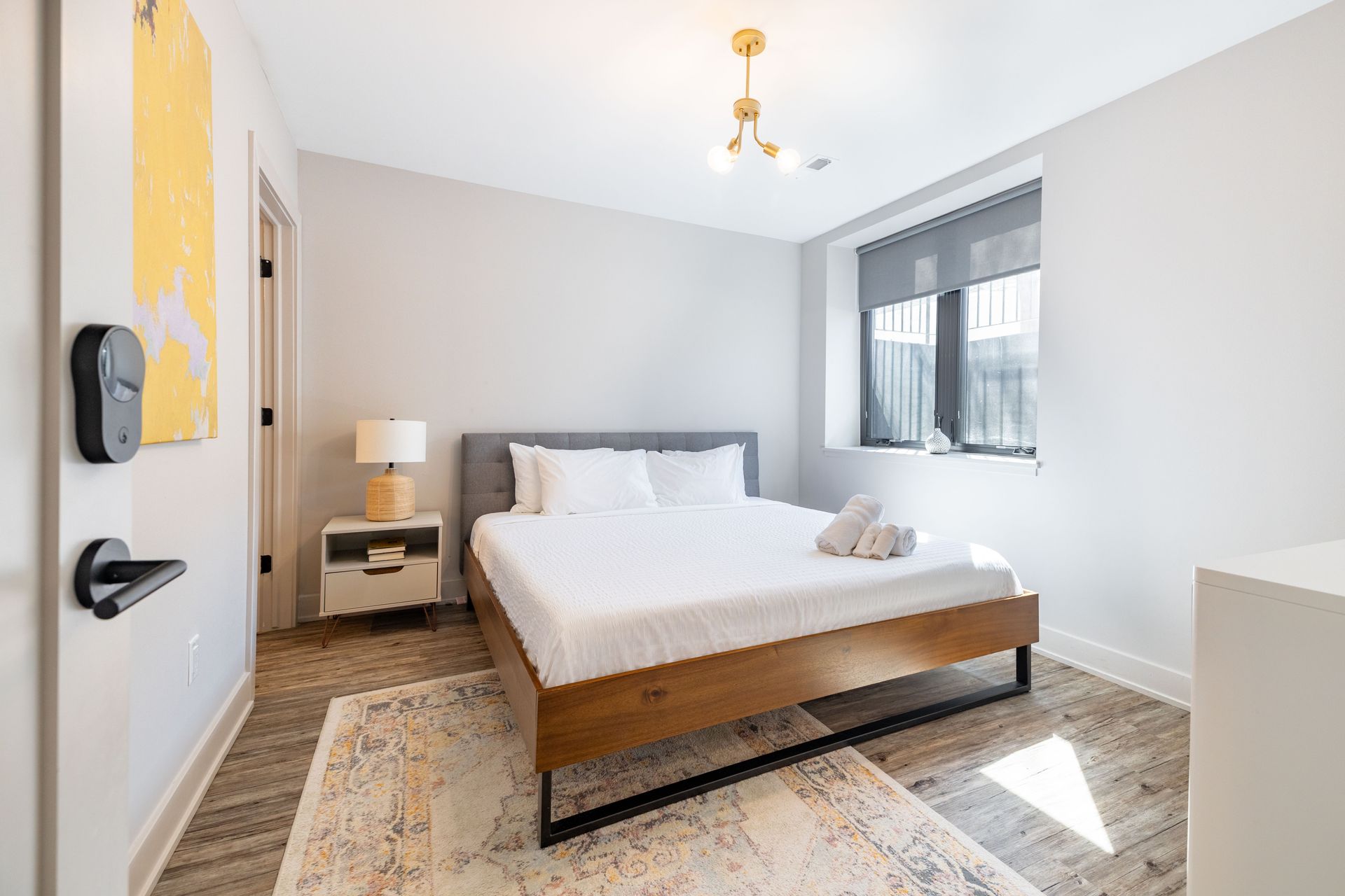 Bright modern bedroom with a white bed, patterned rug, nightstand, and large window.