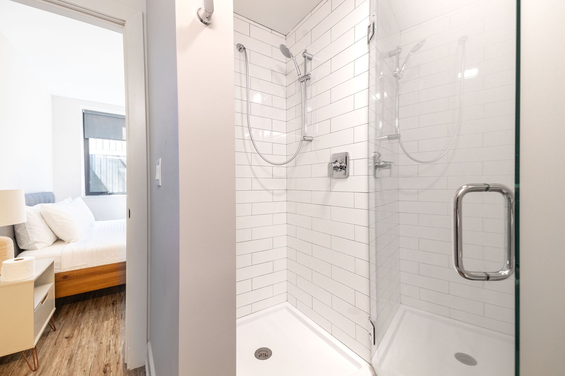 White tiled walk-in shower with glass door beside a bright bedroom seating area