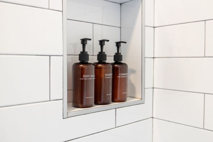 Three brown pump bottles on a recessed bathroom shelf with white tile walls.