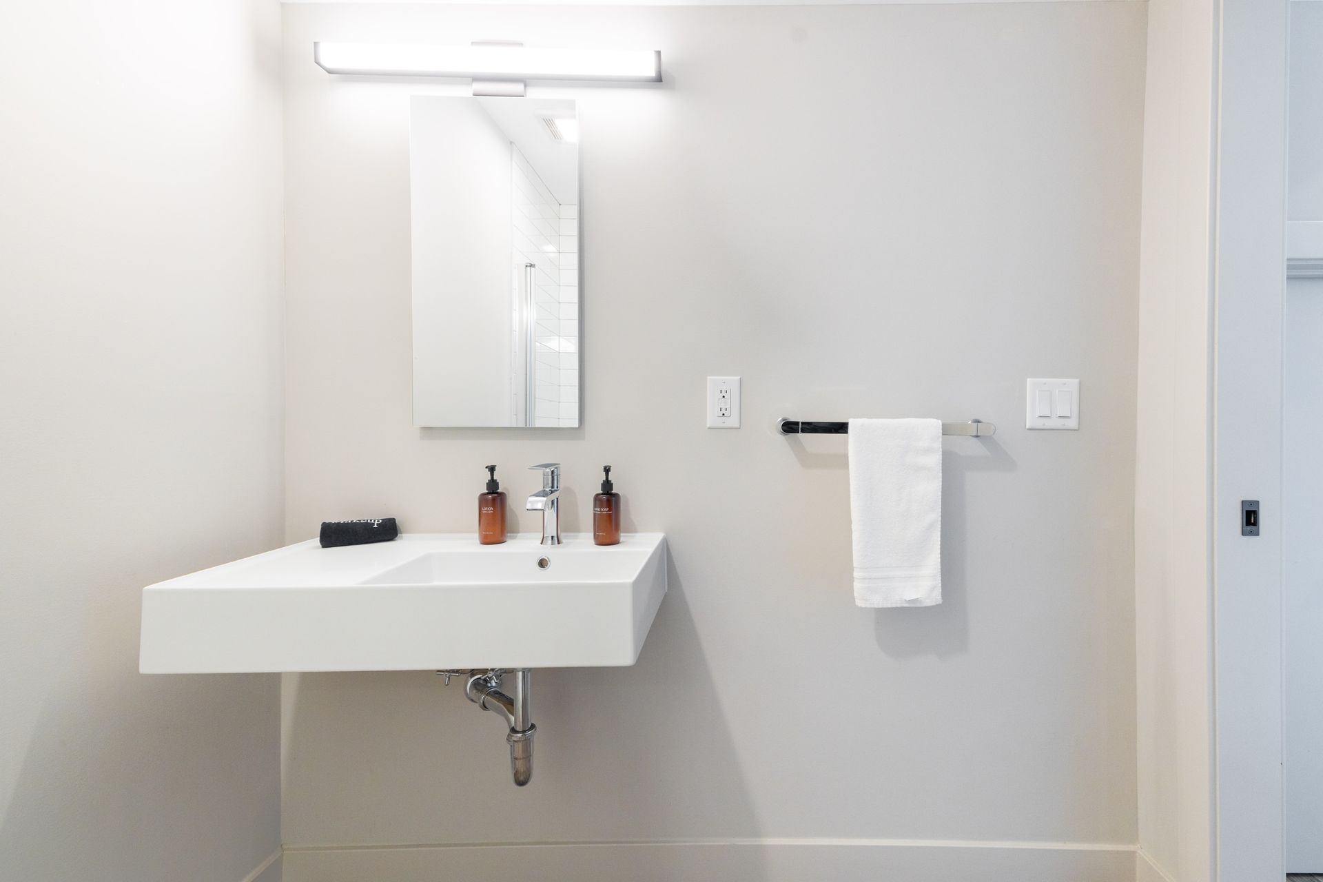 Minimal white bathroom with wall-mounted sink, mirror, light, towel bar, and toiletries