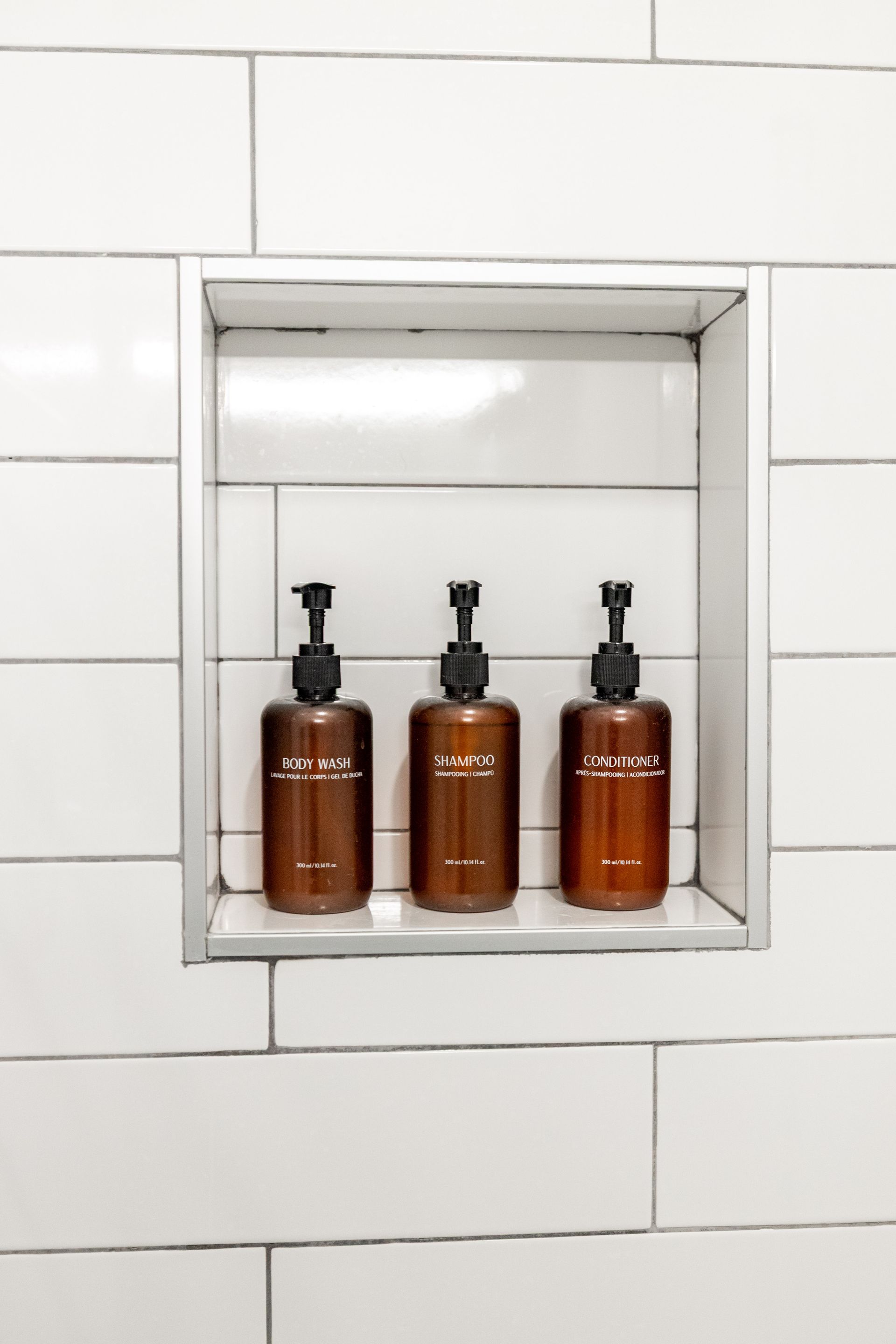 Three brown pump bottles on a built-in shelf in a white tiled shower niche