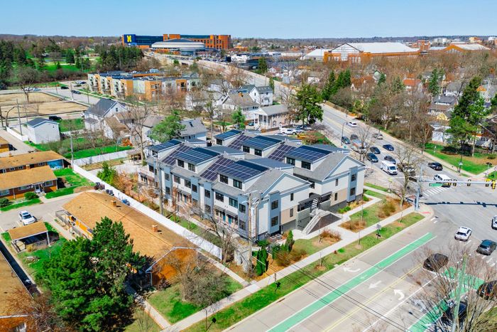 Aerial view of a modern apartment complex beside a road, trees, and nearby buildings