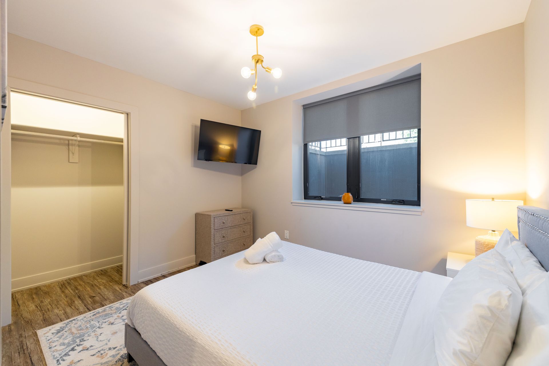 Modern bedroom with a made bed, wall-mounted TV, window, and open closet closet.