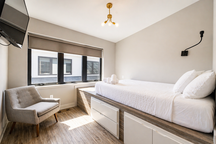 Bright bedroom with a single bed, gray chair, and large windows with blinds