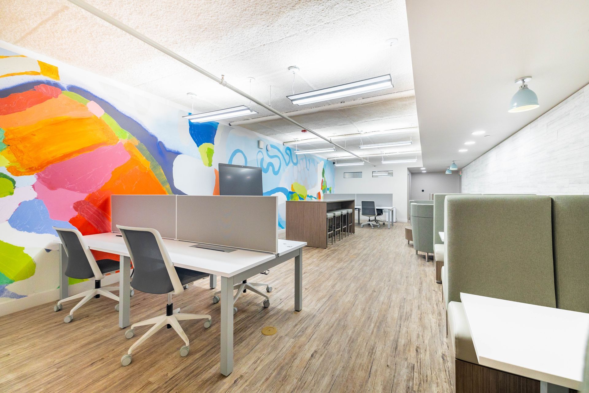 Modern open-plan office with colorful mural, desks, white chairs, and booth seating