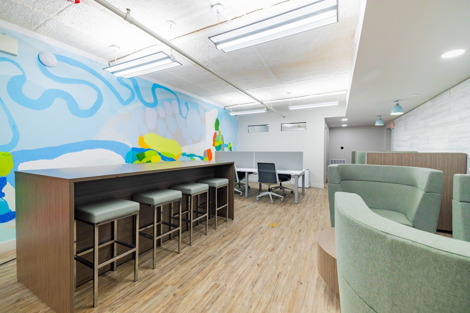 Modern lounge with bar stools, pastel mural wall, and green chairs in a bright office space