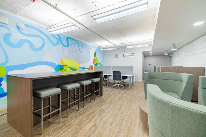 Modern office lounge with bar stools, green chairs, wood flooring, and a colorful mural wall