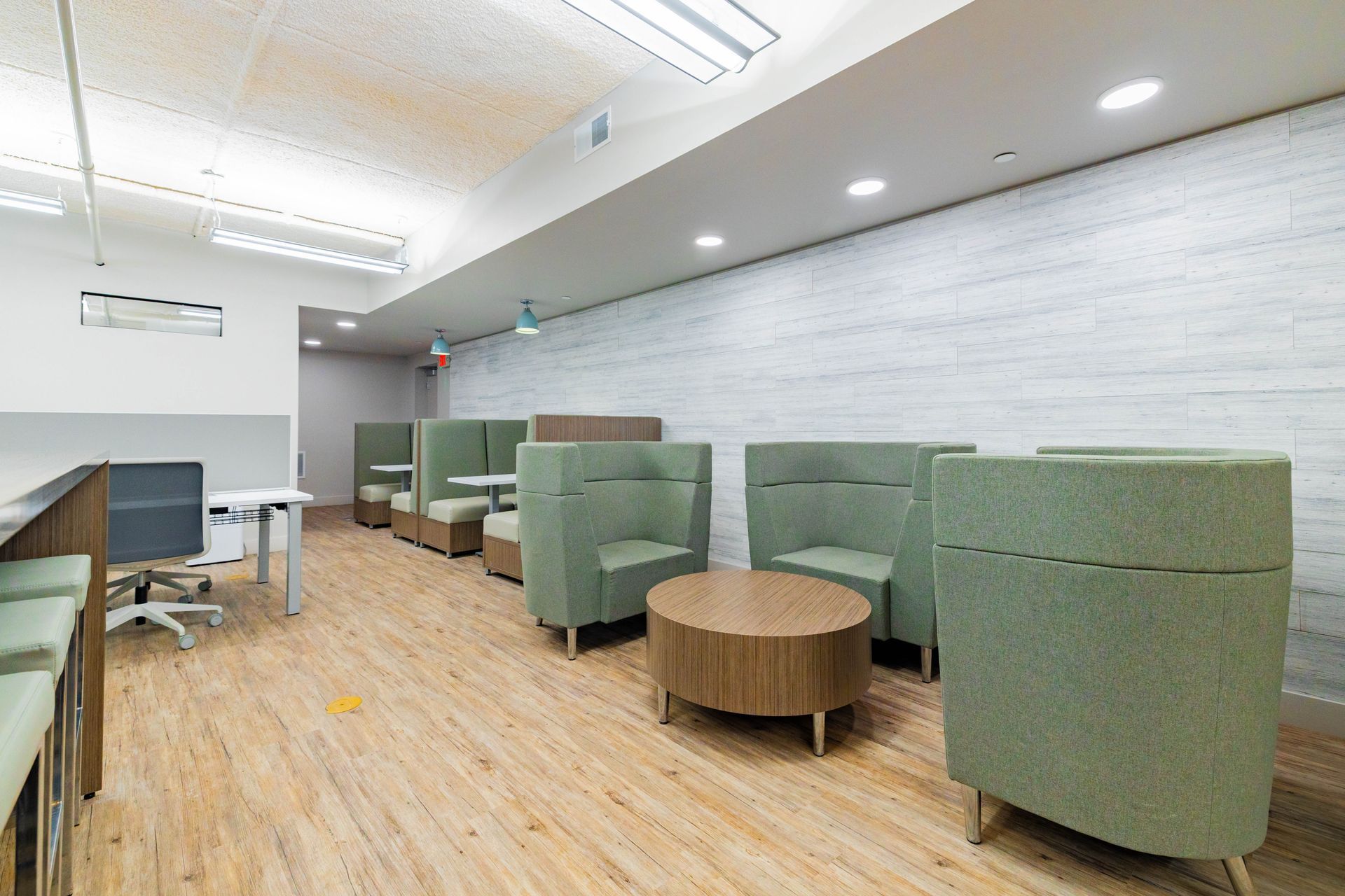 Modern lounge with green armchairs, round tables, wood floor, and a projector screen in a bright room
