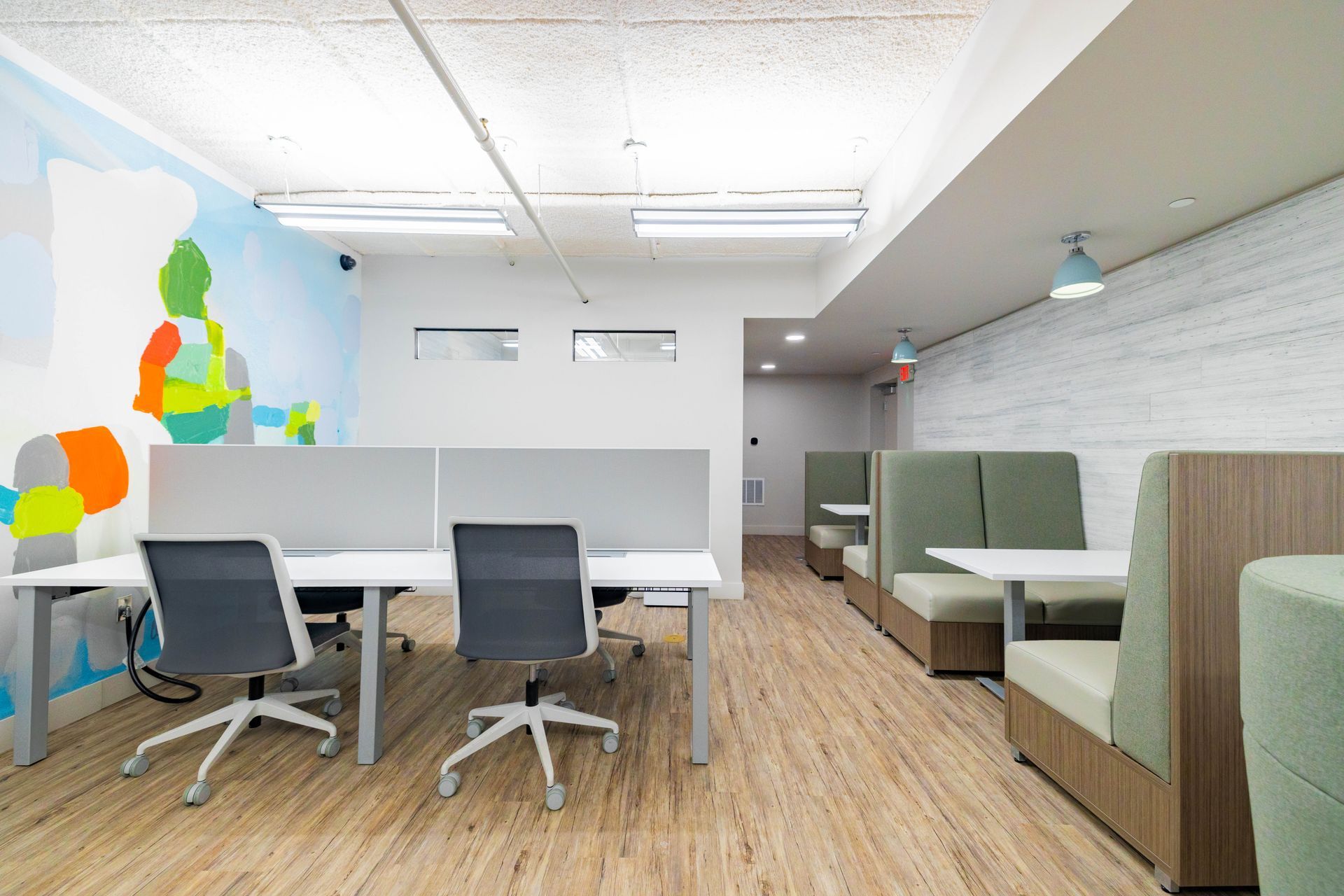 Modern office lounge with cubicles, gray chairs, green booths, wood floor, and colorful wall art