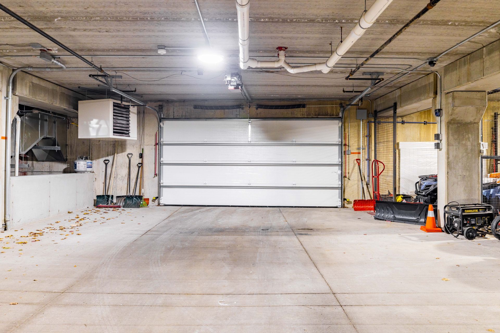 Empty concrete garage with a white roll-up door, exposed ceiling pipes, and tools along the walls