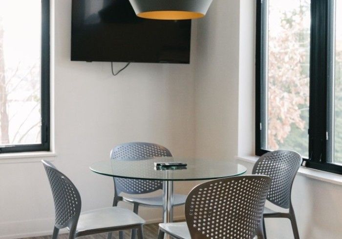 A small dining area with a glass table, four gray chairs, a mounted TV, and windows.