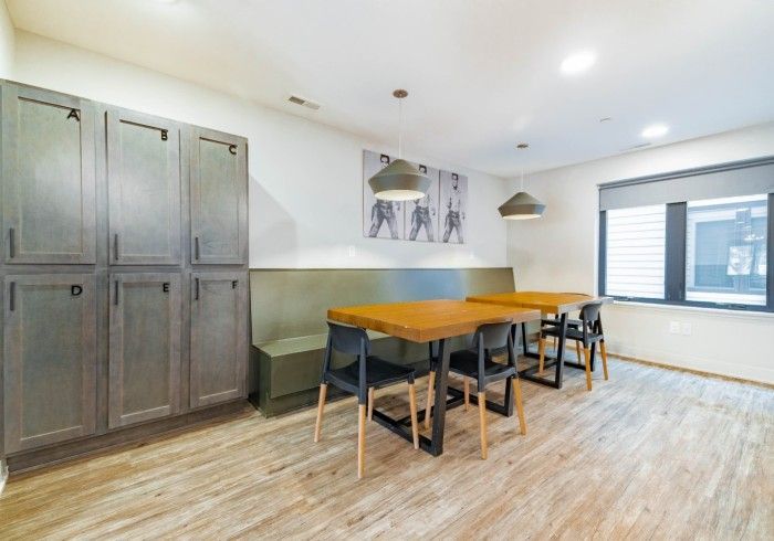 A dining area featuring a long olive-green booth, two wooden tables, black chairs, gray storage lockers, and pendant lights.