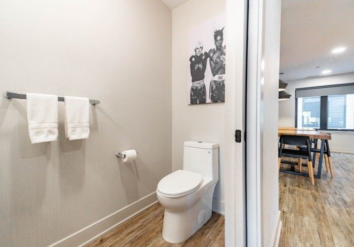A small, minimalist bathroom with a white toilet, two white hand towels on a wall rack, and a black-and-white art print.