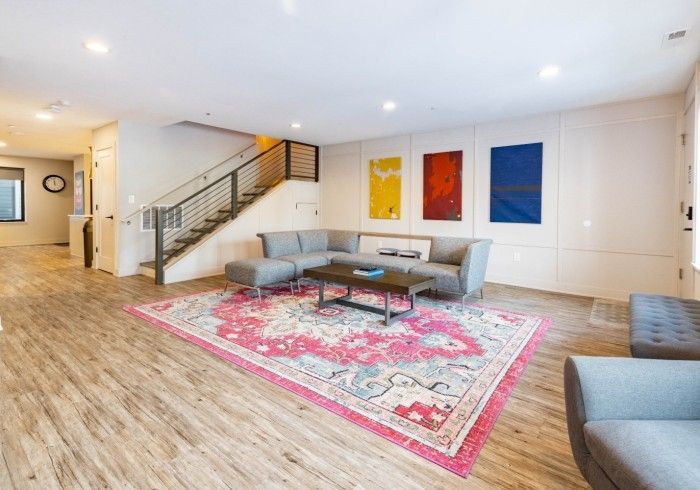 A modern living room with gray furniture, a colorful patterned rug, and three abstract wall art panels near a staircase.