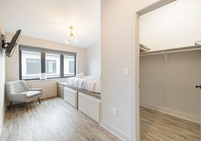 Bedroom with bed, chair, window, closet, and mounted TV. White walls, wood-look floor.