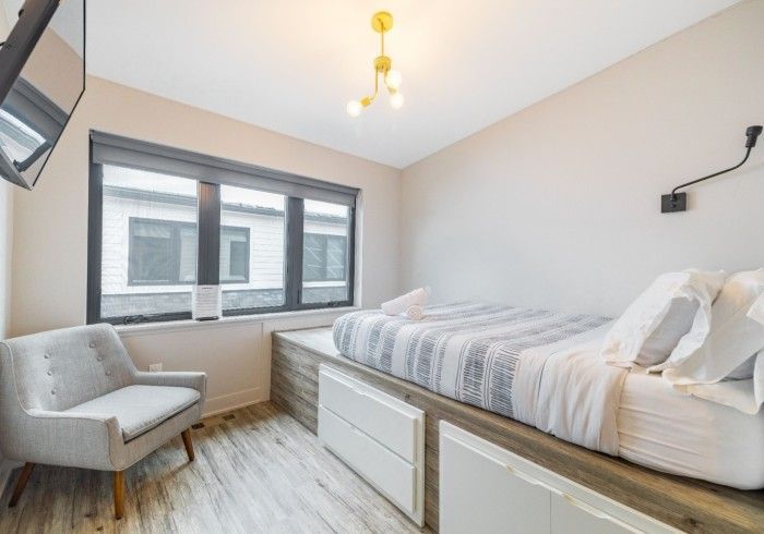 A modern bedroom with a grey armchair, a platform bed with integrated white storage drawers, and a window with grey trim.