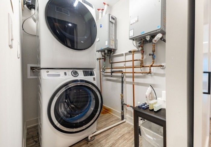 A stacked washer and dryer in a small utility room with exposed copper pipes and a wall-mounted water heater.