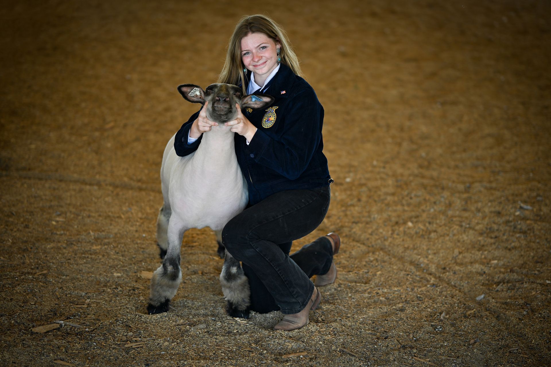 A young person in a blue jacket kneels, embracing a white sheep in a dirt-floored arena.