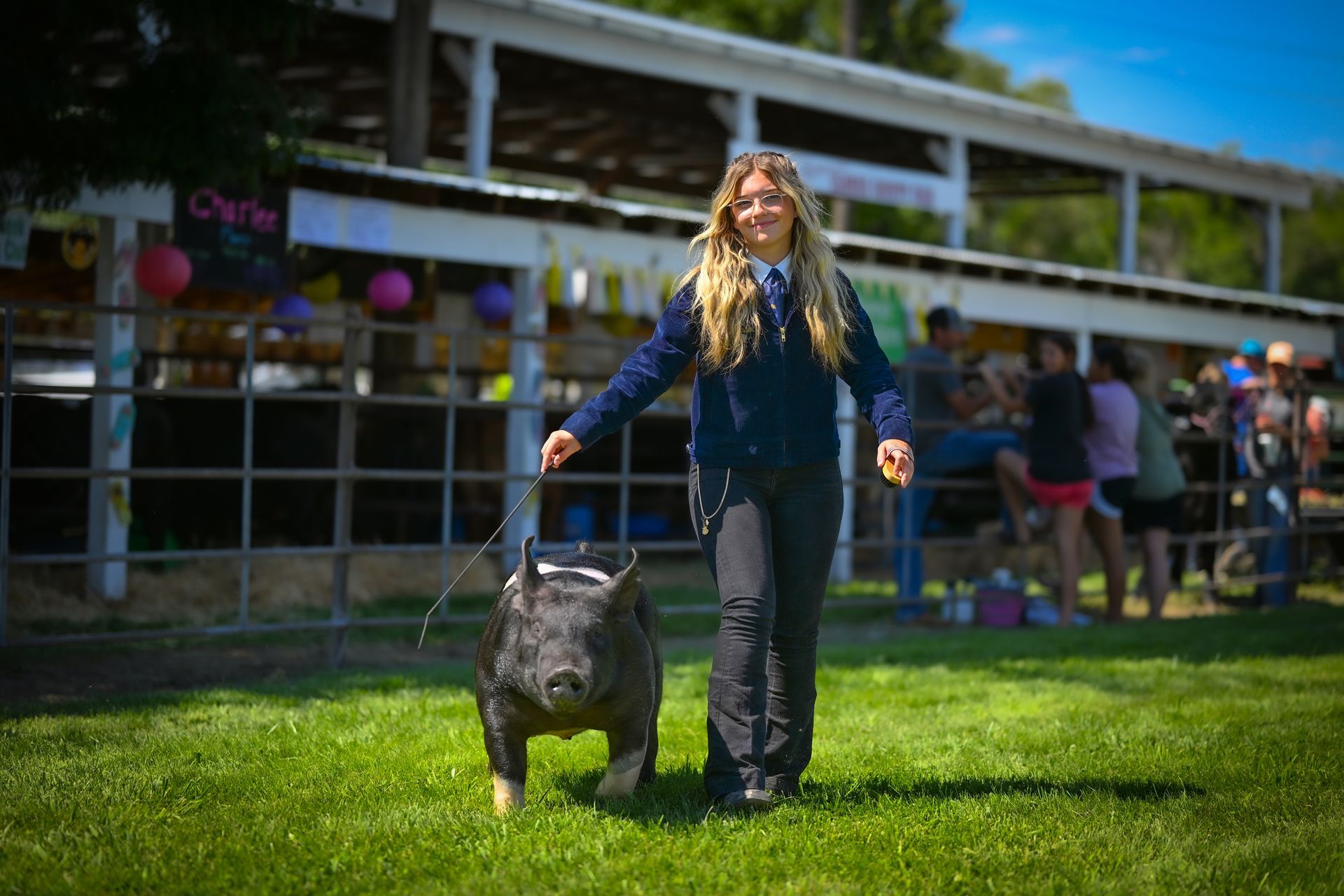 Girl in navy jacket leads a black goat on a leash in an outdoor fair setting.