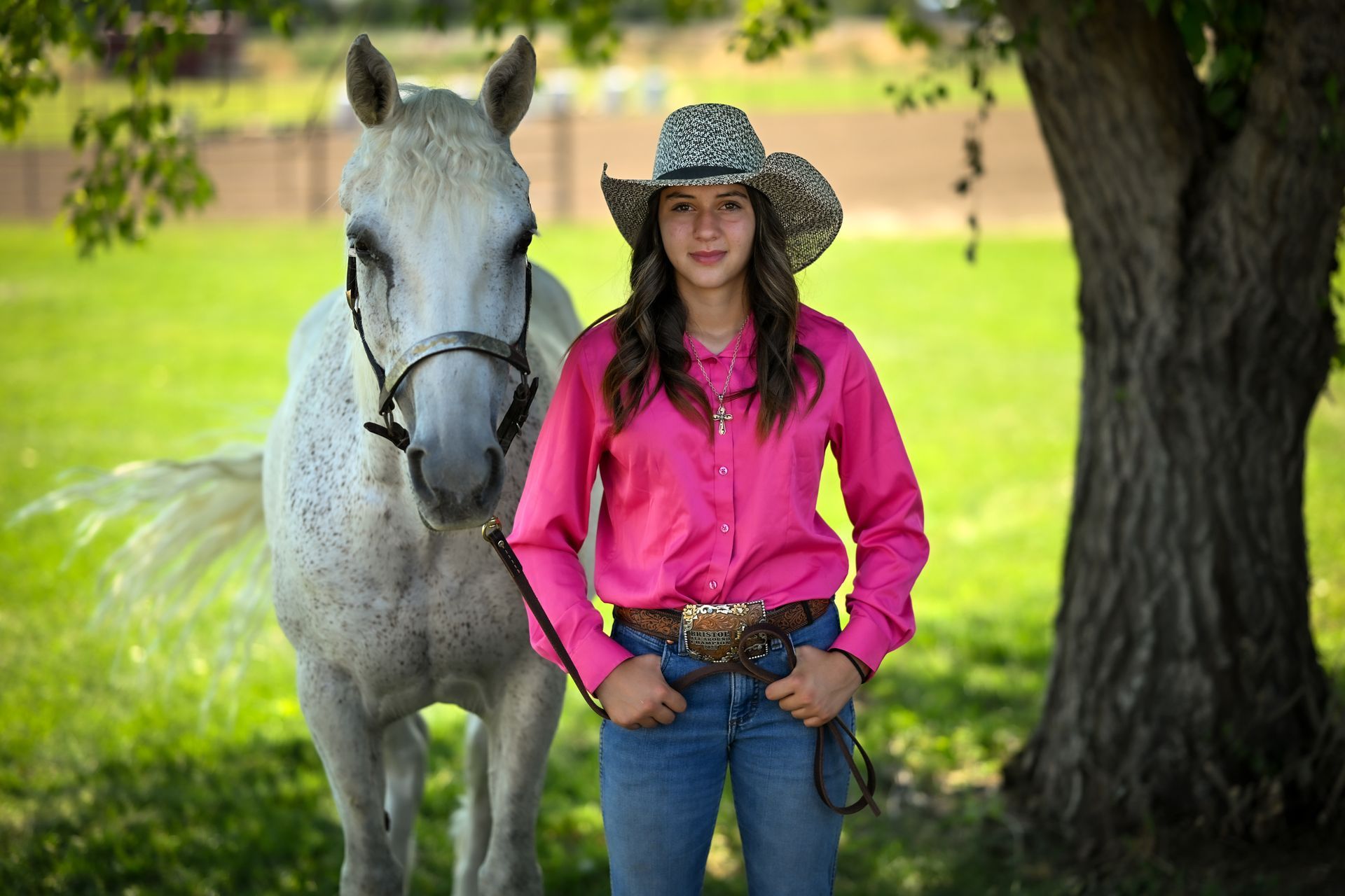 Woman in cowboy hat and pink shirt stands with a white horse in a grassy field.