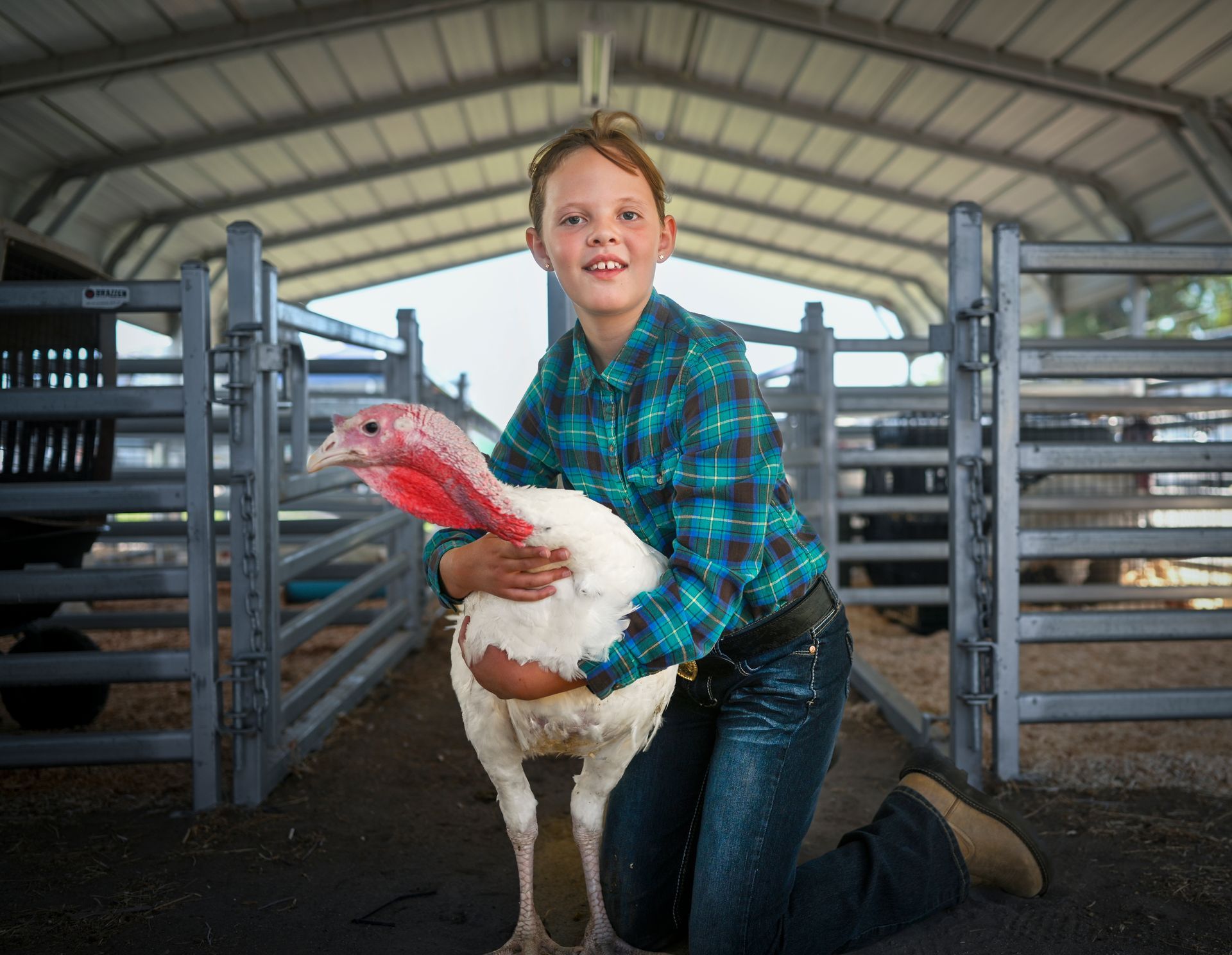 Girl kneeling, hugging a white turkey in a barn. Smiling, wearing plaid shirt and jeans.