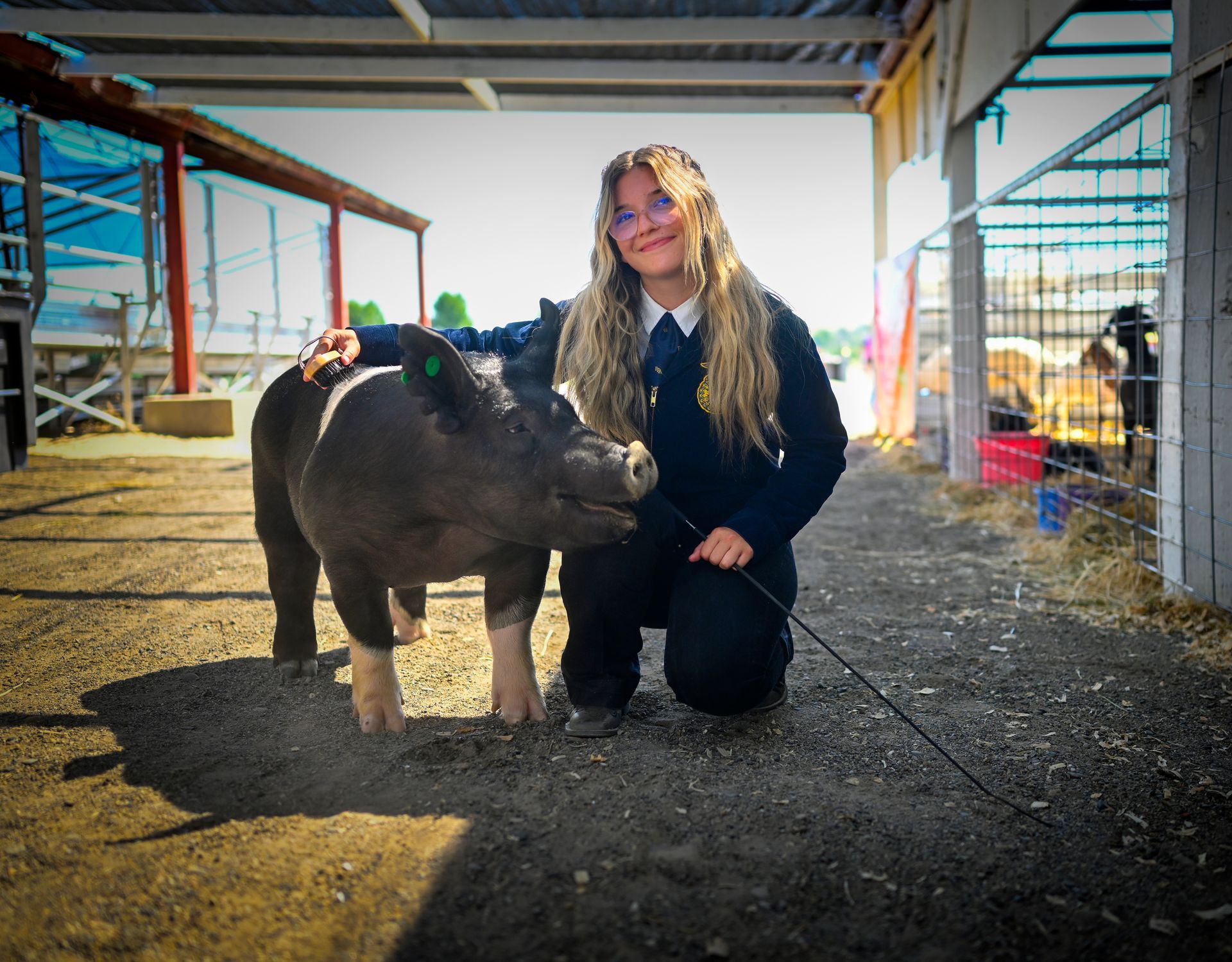 A smiling person kneels with a pig at an outdoor farm. Both look towards the camera.