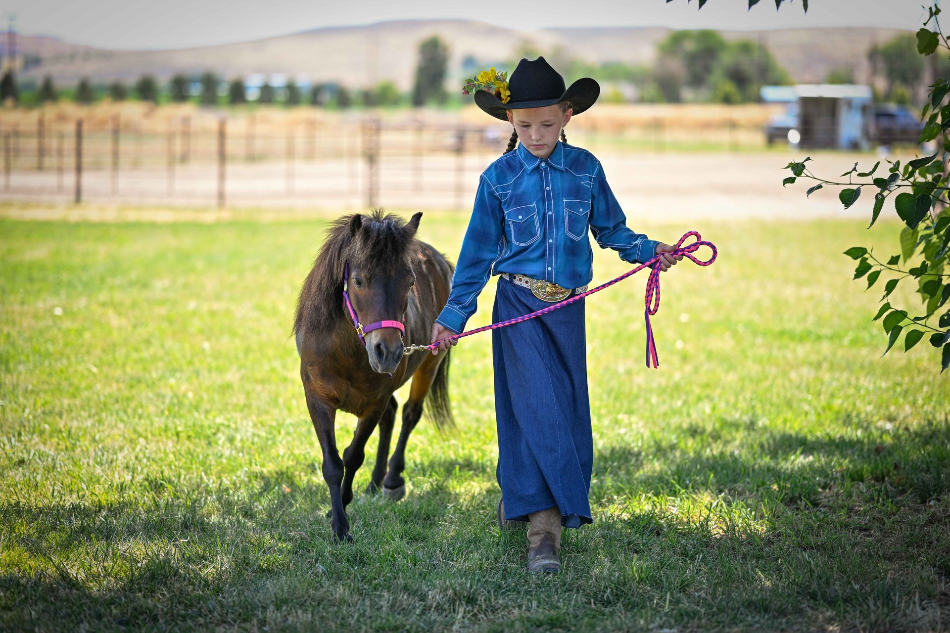 A young boy in a cowboy hat is walking a pony in a field.